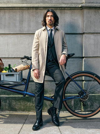 En man klädd i en kostym i skandinavisk design och en vattentät trenchcoat i sandkornmönster lutar sig mot en lastcykel med matvaror.