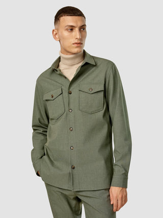 En man klädd i en Essential Overshirt i färgen Nightfall Green Melange över en beige polotröja, med skandinavisk design.