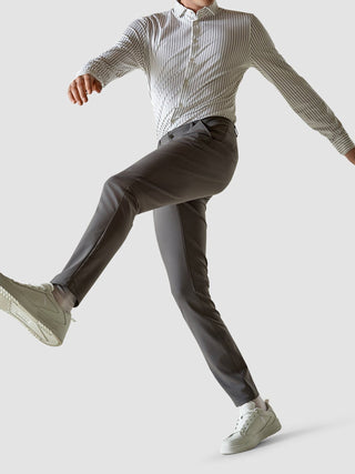 En man i en randig skjorta med knappar och ett par grå Essential Pants Slim, avbildad i en pose mitt i ett steg.