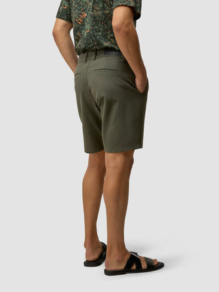 En person i en mönstrad skjorta, GEN2™-shorts i dämpad grön färg med händerna i fickorna och svarta sandaler.