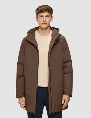 En man bär en brun parka av märket Utility Jacket Saddle Brown över en ljus tröja och mörka byxor.
