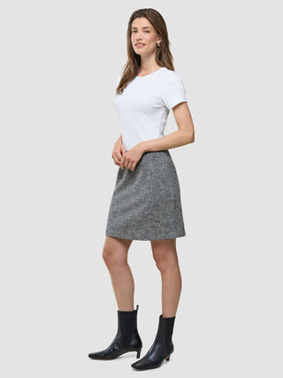 En kvinna bär en vit t-shirt, en kort kjol från Women Ashryn i mönstret ”Domino Herringbone” samt svarta ankelstövlar framför en enfärgad bakgrund.