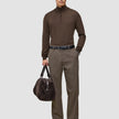 En man klädd i en brun tröja med kvartslång dragkedja och byxor av modellen Atelier Pants Relaxed Dark Beige, som håller i en mörk väska.