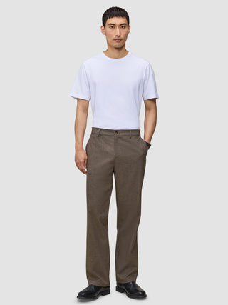 En man i en vit t-shirt, byxor av modellen Atelier Pants Relaxed i mörkbeige och svarta läderskor.