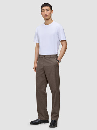 En man i en vit t-shirt, byxor av modellen Atelier Pants Relaxed i mörkbeige och svarta kostymskor.