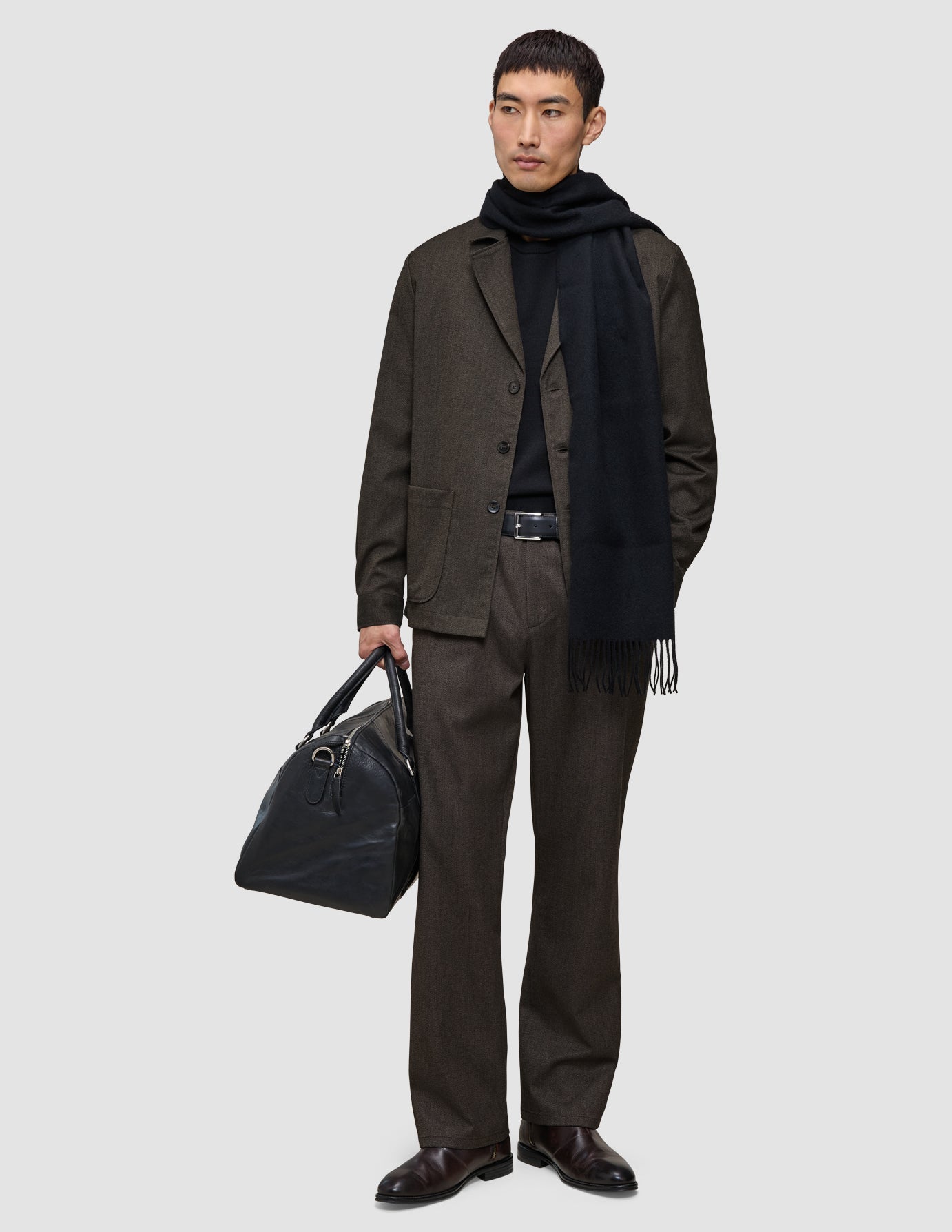 En man klädd i ett par avslappnade byxor från Atelier Pants Relaxed i mörkbrunt och en svart halsduk bär en svart läderväska.
