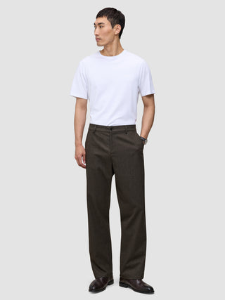 En person i en vit t-shirt, byxor av modellen Atelier Pants Relaxed i mörkbrunt och mörka läderskor.