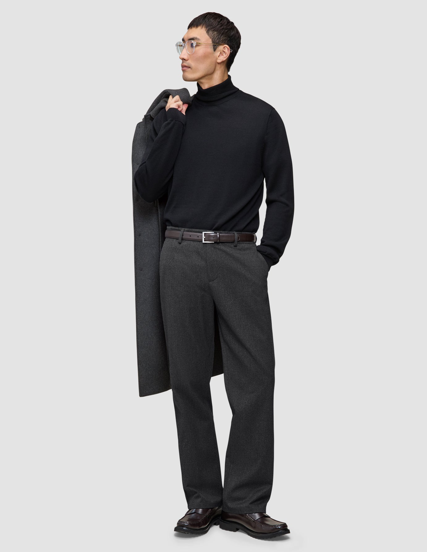 En man i svart polotröja och ett par Atelier Pants Relaxed i mörkgrått med avslappnad passform, med en kappa över axeln.