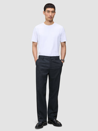 En man bär en vit T-shirt med rund hals och byxor av modellen Atelier Pants Relaxed Dark Navy i bruten twill.