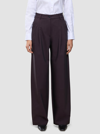 En person bär en vit skjorta med knappar och dambyxorna Avira Pants Wide i färgen Burgundy Berry, som har en avslappnad passform med vida ben.