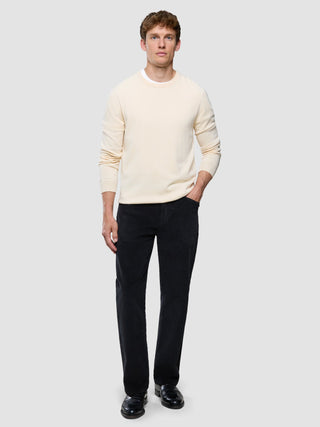 En man klädd i en krämfärgad tröja med rund hals, svarta corduroybyxor i avslappnad passform och svarta loafers.