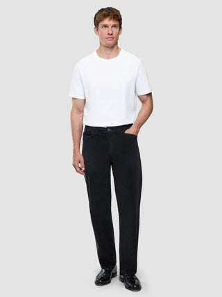 En man i en vit t-shirt och svarta corduroybyxor i avslappnad passform står framför en ljus bakgrund.