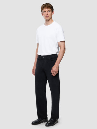 En man klädd i en vit t-shirt och svarta corduroybyxor i avslappnad passform står framför en enfärgad bakgrund.