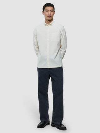 En man klädd i en benvit corduroyskjorta, mörka jeans och svarta skor, som står med händerna bakom ryggen.