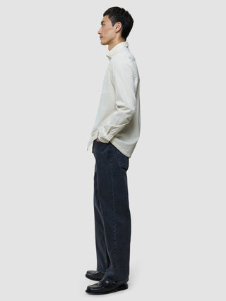 En man klädd i en benvit corduroyskjorta, mörka jeans och svarta loafers, avbildad i profil.