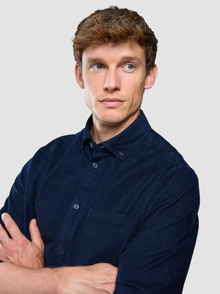 En man med ljust hår, klädd i en mörkblå corduroy-skjorta, står med armarna i kors.