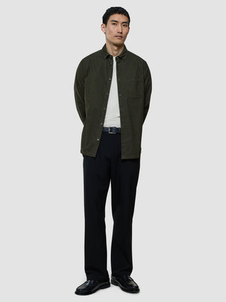 En man står där klädd i en öppen corduroyskjorta i färgen Nightfall Green över en vit t-shirt, svarta byxor och svarta loafers.