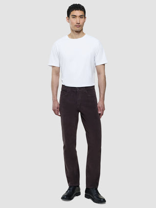En man i en vit T-shirt med rund hals och corduroybyxor i färgen Deep Espresso, som står framför en enfärgad bakgrund.