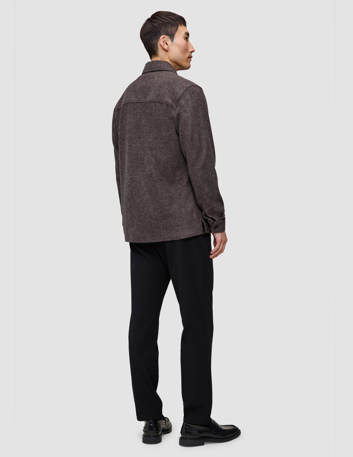 En person sedd bakifrån, klädd i en dubbellagad ylleöverskjorta i sadelbrunt, svarta byxor och svarta loafers.