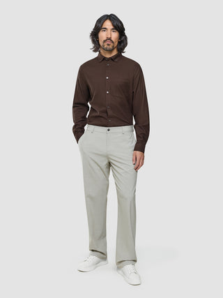 En man med långt mörkt hår, klädd i en brun skjorta med knappar, Ease Pants Relaxed Fit Sahara och vita sneakers.
