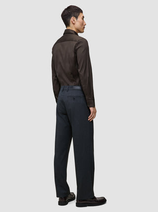 En man sedd bakifrån, klädd i en mörkbrun, figursydd skjorta med hög krage, kolgrå byxor och mörka loafers.