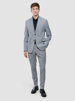 En man klädd i en Essential Blazer Regular Ash Twill tillsammans med en svart skjorta och svarta loafers, vilket återspeglar en minimalistisk design.