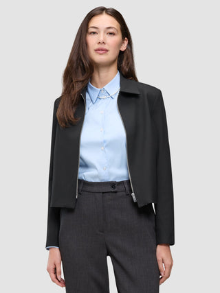 En kvinna bär en svart Essential Short Zip Jacket över en ljusblå skjorta med knappar och grå byxor.