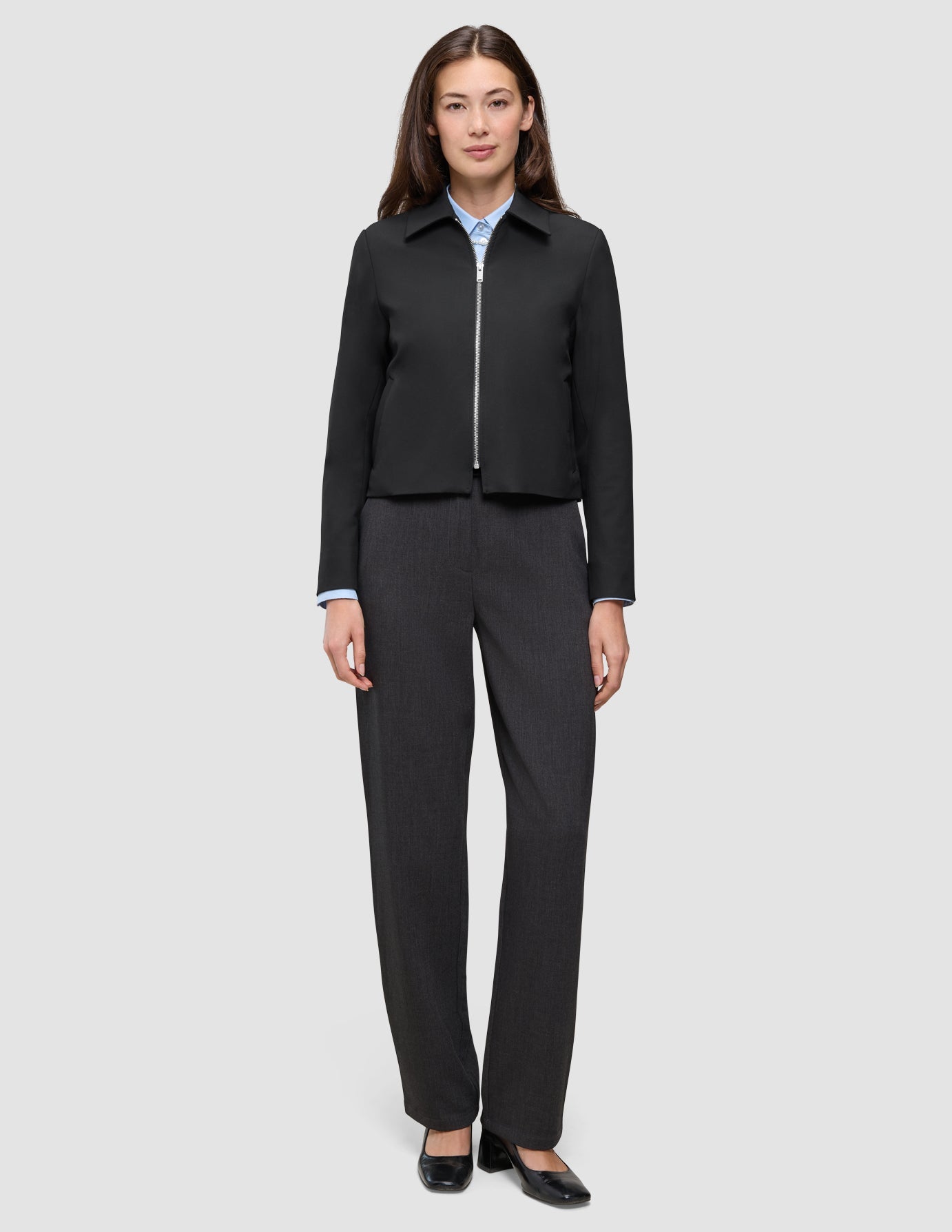 En kvinna som bär en svart Essential Short Zip Jacket över en ljusblå skjorta med krage och mörkgrå byxor.