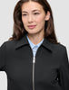 En person som bär en minimalistisk Essential Short Zip Jacket Black över en ljusblå skjorta med knappar.