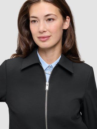 En person som bär en minimalistisk Essential Short Zip Jacket Black över en ljusblå skjorta med knappar.