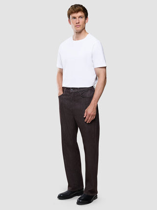 En man klädd i en vit t-shirt, ett par Marco Jeans i mörkbrunt med avslappnad passform och svarta skor.