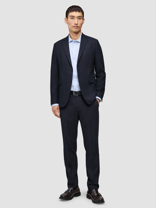 En man klädd i en blazer av modellen Motion Blazer Regular i färgen Midnight Blue och matchande byxor, en ljusblå skjorta och mörka loafers.