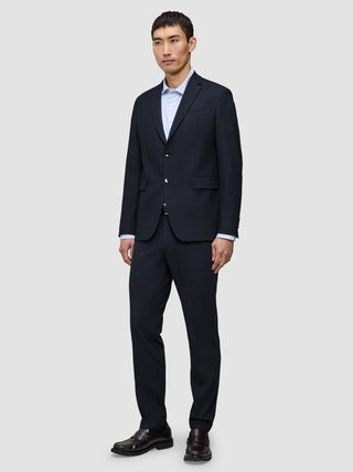 En man klädd i en kostym från Motion Blazer i storlek Regular i färgen Midnight Blue och matchande byxor, med en ljusblå skjorta och bruna loafers.