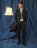 En man klädd i byxor av modellen Motion Suit Pants Relaxed Cognac, en vit skjorta och en slips står bredvid en golvlampa.