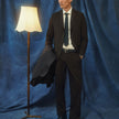En man klädd i byxor av modellen Motion Suit Pants Relaxed Cognac, en vit skjorta och en slips står bredvid en golvlampa.