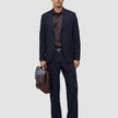 En man klädd i mörkblå byxor i modell Midnight Blue Motion Suit med avslappnad passform, en mörk skjorta och bruna loafers håller i en läderportfölj.