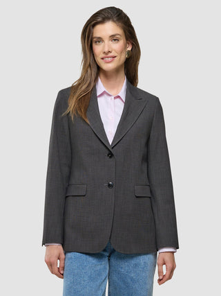 En leende kvinna klädd i en Women Nara Blazer Cool Brown, över en ljusrosa skjorta med knappar och blå jeans.