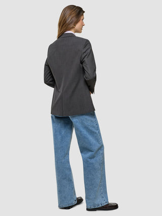 En kvinna klädd i en ”Women Nara Blazer Cool Brown”, blå jeans och mörka loafers står med ryggen mot betraktaren och förkroppsligar minimalistisk design.