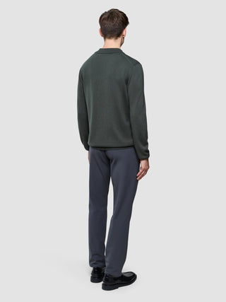 En man sedd bakifrån, klädd i en Seamless Core Knit långärmad polotröja i färgen Green Night, grå byxor och svarta skor.