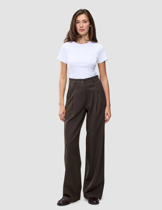 En person bär en vit t-shirt och byxorna Serene Pants Wide Walnut Herringbone: bruna byxor med hög midja och vida ben.
