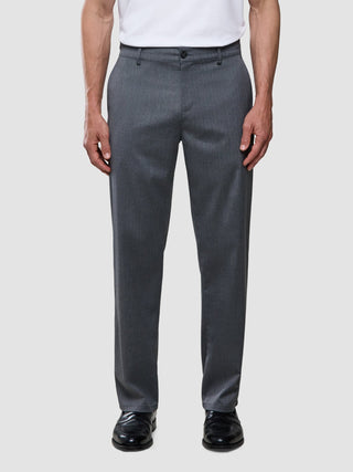 En man bär en vit t-shirt, byxor av modellen ”Siena Pants Relaxed Fit Herringbone Grey” och svarta skor.