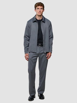 En man klädd i en grå jacka med dragkedja över en svart skjorta och byxor av modellen ”Siena Pants Relaxed Fit Herringbone Grey”.