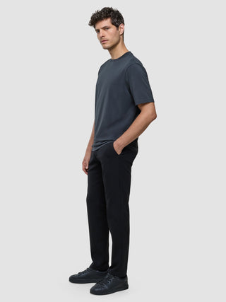 En man klädd i en Supima-T-shirt i Box Fit-modell i mörkgrått, svarta byxor och sneakers står framför en enkel skandinavisk bakgrund.