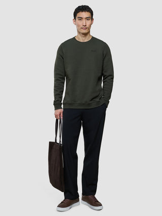 En man bär en grön tröja med rund hals från Sweat Crewneck och svarta byxor, och håller i en mörkbrun tygväska.
