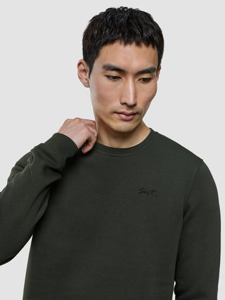 En man klädd i en grön tröja med rund hals från märket Sweat Crewneck Green Night, med bokstäverna ”Snt.” broderade på bröstet.