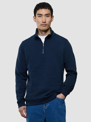 En man klädd i en mörkblå tröja med halv dragkedja och blå jeans.