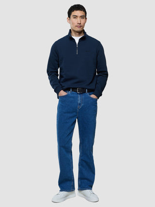 En man klädd i en mörkblå tröja med halv dragkedja, blå jeans och vita sneakers.