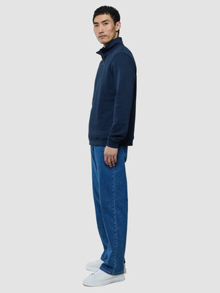 En man i profil klädd i en mörkblå tröja med halv dragkedja, blå jeans och vita sneakers.
