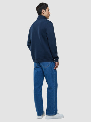 En man sedd bakifrån, klädd i en mörkblå tröja med halv dragkedja, blå jeans och vita sneakers.