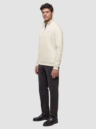 En man klädd i en offwhite tröja med halv dragkedja, mörka byxor och loafers.